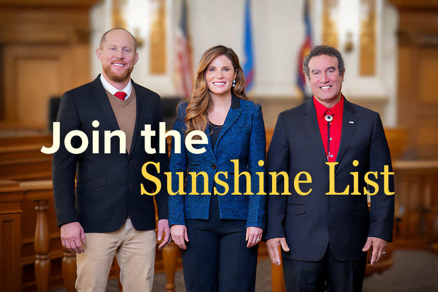 The Mesa County Board of Commissioners — Cody Davis, Bobbie Daniel and JJ Fletcher — stand side by side in the public hearing room, wearing business attire and smiling, with flags and wood-paneled seating in the background. The text “join the sunshine list” is in yellow on top of the image.