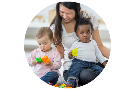 A woman sitting on the floor with two young toddlers. One toddler sits in her lap holding a yellow plastic toy block, while the other sits beside her wearing a pink shirt, playing with green and orange blocks. The woman looks down lovingly at the children in a bright, play-centered environment.